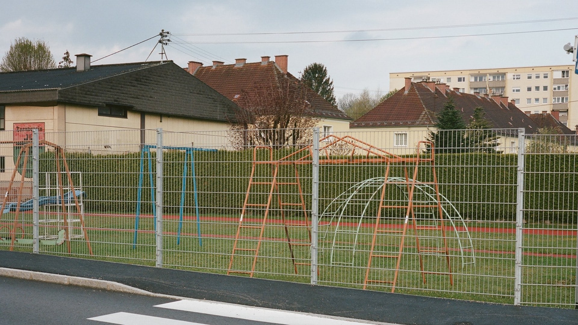 Bunte Spielgeräte auf einem Schulhof, eingezäunt, bei bewölktem Himmel in Eibiswald.