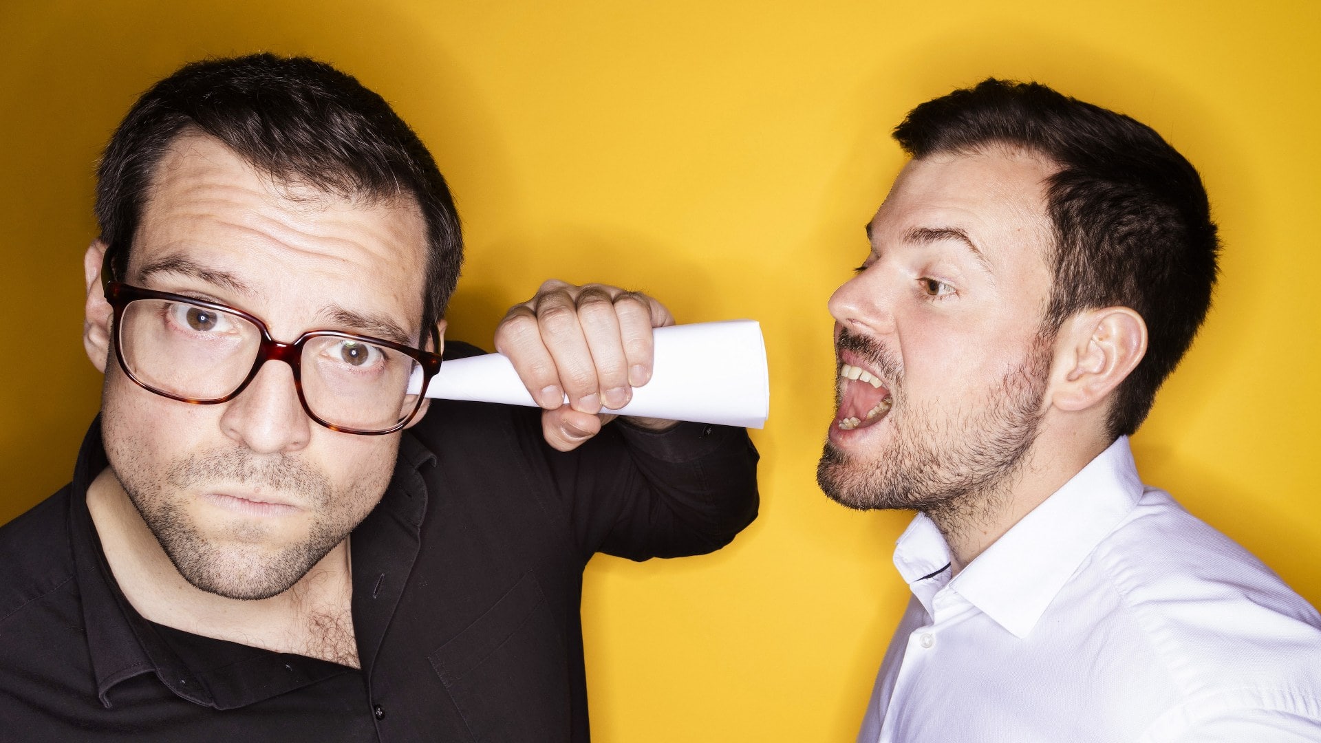 Ein Mann hält eine Papierrolle an seinem Kopf, während ein anderer Mann laut spricht, vor einem gelb.